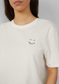 T-shirt en coton blanc à manches courtes, avec un petit motif de texte imprimé sur le côté gauche de la poitrine indiquant "c'est ce que c'est."