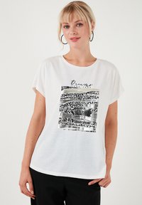 Hvid bomuld t-shirt med rullede ærmer, prydet med et sort og metallisk tryk med ordet "Drømme" og abstrakte mønstre. Blød tekstur.