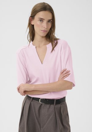 Mujer joven con cabello castaño que lleva una camiseta rosa claro con cuello en V y pantalones grises de rayas finas de cintura alta con un cinturón negro, con los brazos cruzados.