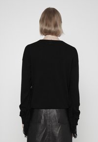 Pull en maille noire à manches longues, avec des franges sur les côtés. Le dos présente un col rond simple.