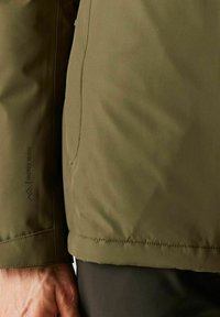 Primer plano de una persona con una chaqueta aislante color verde oliva con el texto "Isotex 15000" en la manga, mostrando detalles de costura y la mano.