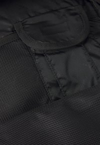 Črna tkaninska torba z mrežnim žepom, gladko teksturo in ojačanim robom. Zapiranje vključuje majhno pokrovko in zanko za nošenje.