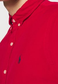 Camicia rossa con colletto e patta con bottoni, caratterizzata da un piccolo logo nero sul lato sinistro. Il tessuto sembra avere una trama a costine.