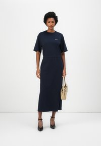 Ναυτικά μπλε, μακρύ maxi φόρεμα με χαλαρή εφαρμογή, κοντά μανίκια και ελαστική μέση. Συμπληρώνει μια γυναίκα που κρατά μια μπεζ τσάντα και φορά μαύρες γόβες.