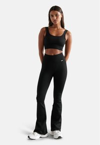 Schwarzes, geripptes Cropped Top und hochtaillierte, ausladende Leggings, beide mit einer strukturierten Oberfläche. Akzentuiert mit einem kleinen Logo am Taillenbund. Darunter weiße Sneaker.