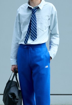 Homme portant une chemise blanche, une cravate rayée bleue nouée de manière lâche, un pantalon de survêtement bleu vif, et tenant un sac de sport noir.