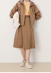 Femme portant une veste marron et une jupe avec une chemise rayée orange, portant un sac à dos blanc et des baskets beiges sur fond blanc.