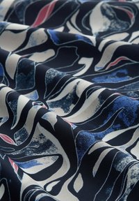Tessuto patterned con motivi a spirale in blu navy, bianco e rosso. La texture è liscia con una leggera lucentezza, creando profondità e contrasto.