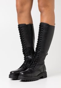 Mustat polvipituiset saappaat, jotka on valmistettu sileästä nahasta. Niissä on edessä nauhoitus, kestävä kumipohja ja sivu-zip, joka helpottaa pukemista.