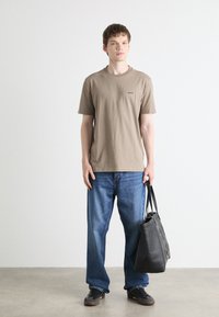 Beige bomull T-skjorte med "BOSS"-logo, avslappet passform, parret med mørkeblå jeans og holder en svart lær tote bag. Uformelt fottøy inkludert.
