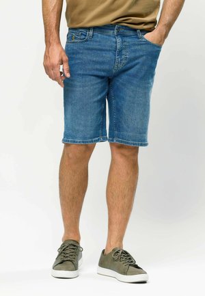 Mand iført blå denimshorts, beige skjorte og olivengrønne sneakers, stående med den ene hånd i lommen mod hvid baggrund.