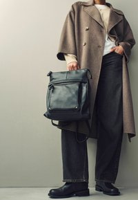 Sac à dos en cuir noir avec poche frontale, porté par une personne en manteau marron et jean noir. Design simple, bretelles robustes et accents métalliques.