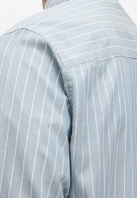 Ανοιχτό μπλε πουκάμισο με λεπτές λευκές κάθετες ρίγες, κατασκευασμένο από μαλακό ύφασμα με ομαλή υφή και σχέδιο κλασικού γιακά.