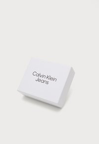 Vit rektangulär låda med en slät yta. Framsidan visar "Calvin Klein Jeans" i svart, modern stil. Lådan är något vinklad för synlighet.