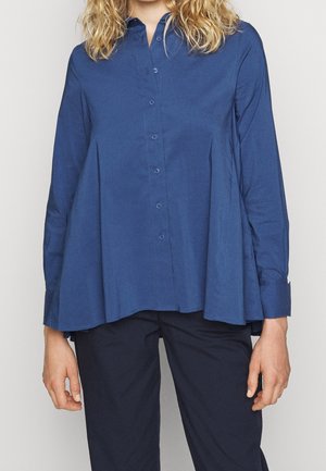 Vrouw draagt een losse, blauwgeknoopte lange mouwen blouse met een donkere broek, staand tegen een effen achtergrond.