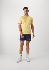 Camiseta deportiva amarilla con detalles en blanco, combinada con pantalones cortos azul marino. Modelo vistiendo zapatillas blancas y calcetas, de pie sobre un fondo neutro.