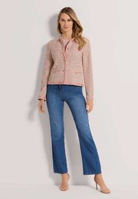 Rosa- und cremefarbener Tweed-Blazer mit goldenen Knöpfen, zwei Fronttaschen und gewellten Kanten. Kombiniert mit ausgestellten blauen Jeans und transparenten Heels.