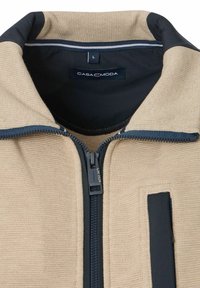 Tans kleurige ritsjacket van een gestructureerde stof, met zwarte accenten bij de kraag en rits, en een klein zakje aan de rechter voorkant.
