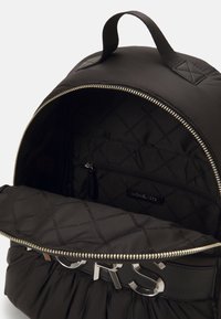Zaino in nylon nero con un interno trapuntato, accessori in metallo argentato e una tasca con zip. Presenta una maniglia superiore e un design arricciato alla base.