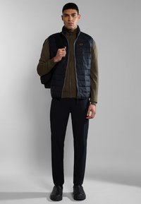 Chaleco acolchado negro sobre una chaqueta verde con cremallera, combinado con pantalones negros ajustados y zapatos oscuros. El chaleco cuenta con un parche de logo circular.