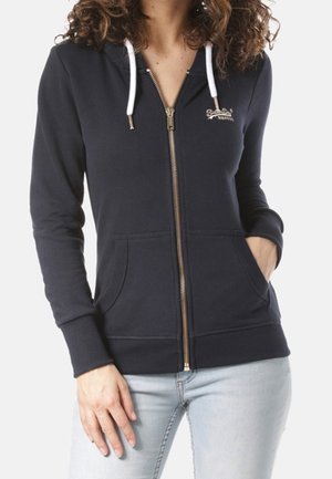 Femme portant un sweat à capuche zippé bleu marine avec des cordons blancs et un jean bleu clair, une main dans la poche et les cheveux bouclés visibles.