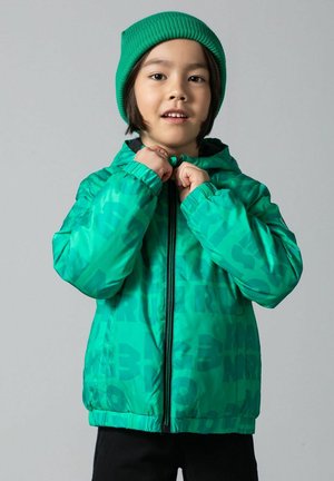 Dziecko w zielonej czapce i zielonej kurtce w wzory, zaciągające zamek kurtki na jednolitym szarym tle.