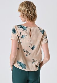 Blusa a maniche corte in beige chiaro con motivi floreali color teal scuro, con dettaglio a oblò sulla schiena e tessuto leggero e liscio.