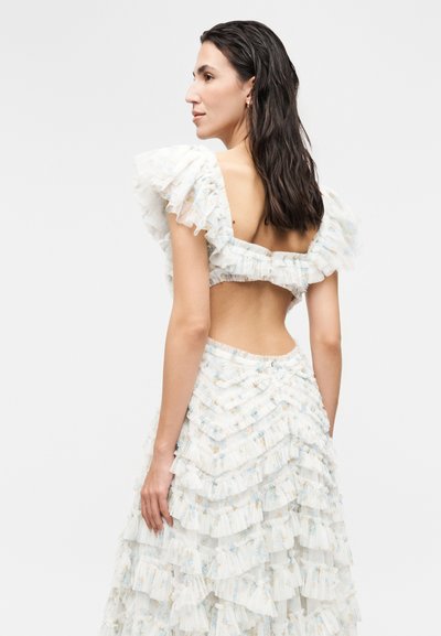 Γυναίκα με σκούρα μαλλιά που φοράει ένα άσπρο φόρεμα με βολάν και floral σχέδια, με ανοιχτή πλάτη, γυρισμένη πλαγίως σε απλό φόντο.