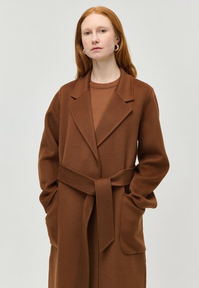 Femeie cu păr roșu și cercei argintii cerc, purtând o haină maro cu centură, reveri mari și mâinile în buzunare, pe fundal simplu.