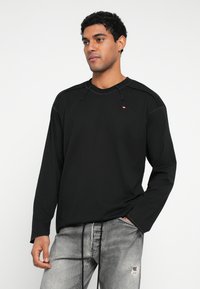 Juoda ilgomis rankovėmis palaidinė su apvalia iškirpte, matomomis siūlėmis ir mažu logotipu ant krūtinės, dėvima su pilkais nudėvėtais džinsais.