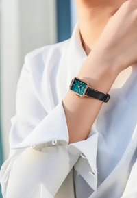 Kvadratformad klocka med en teal ansikte, guld- och silverdetaljer, svart läderarmband, visad på ett handled som bär en vit skjorta.