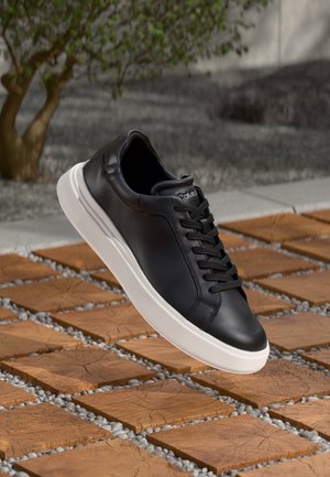 Czarny skórzany sneaker z białą podeszwą zawieszony nad drewnianymi kwadratowymi płytkami i żwirem, na tle liściastego drzewa na świeżym powietrzu.