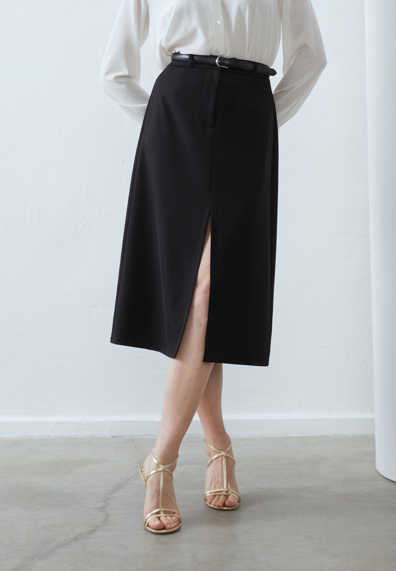Femme portant une jupe midi noire avec une fente devant, un chemisier blanc rentré, une ceinture noire, et des sandales à talons hauts dorées à brides, debout sur un sol en béton.