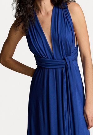 Kobieta w bezrękawną sukienkę o głębokim dekolcie w szpic, w kolorze królewskiego błękitu, z przewiązanym w talii pasem, stoi z jedną ręką zgiętą za plecami.