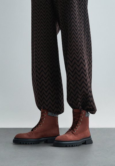 Pruuni ningastabootidel on tekstureeritud ülemine osa, ümar varvas ja mustad kummitald. Asetatud mustrilise kudumise äärega.