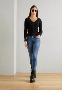 Top de manga larga de canalé negro con cremallera, combinado con jeans ajustados azules y botas de combate negras con cordones, frente a una pared gris claro.