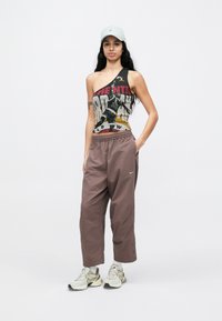 Débardeur asymétrique noir et gris avec impression graphique, accompagné d'un pantalon marron ample, de baskets blanches avec des accents, et d'une casquette gris clair.