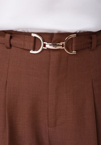 Pantalon taille haute marron en tissu texturé avec une ceinture à boucle argentée brillante ornée d'accents circulaires. Coupe ajustée avec des plis.