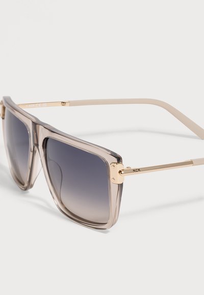 Lunettes de soleil carrées avec verres dégradés gris et montures beige transparent avec des branches fines en métal doré marquées MCM.