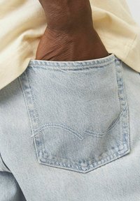 Mörkhyad hand inskjuten i bakfickan på ljusblå denimjeans som bärs med en krämfärgad skjorta.