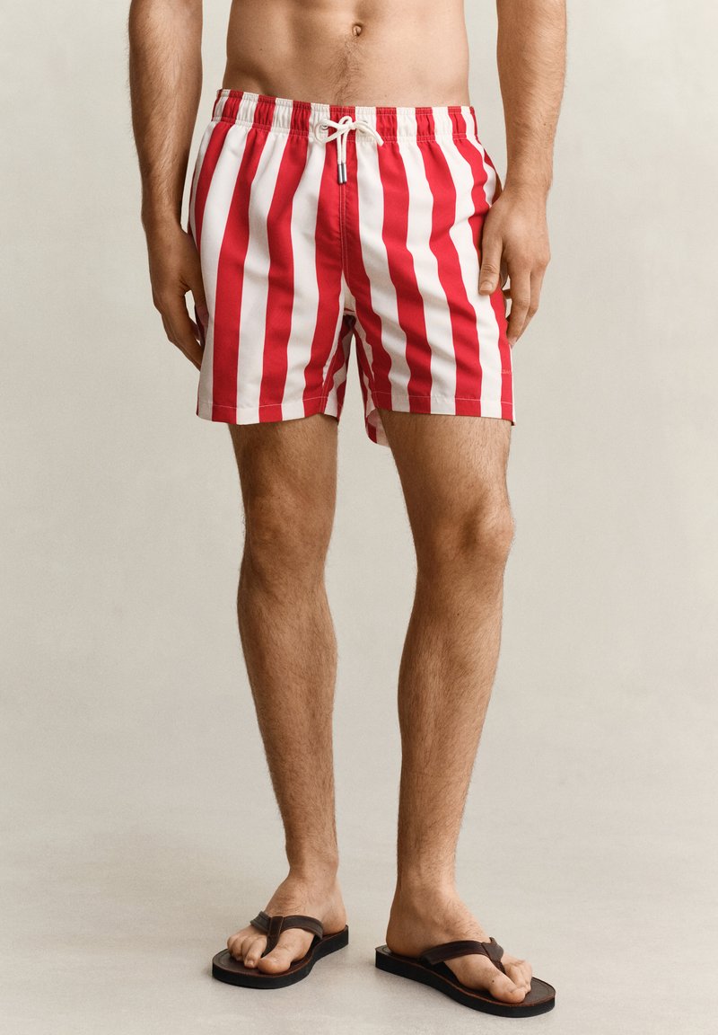 Homme portant un short de bain rayé vertical rouge et blanc et des tongs noires, debout devant un fond clair uni.