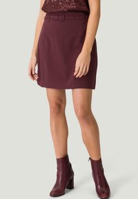 Jupe mini bordeaux avec taille ceinturée. Fabriquée en tissu lisse, elle présente une coupe droite avec un léger éclat, assortie de bottines à la cheville.