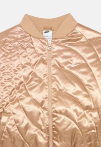 Jaqueta bomber de cetim dourado claro com padrão acolchoado, colarinho canelado, fecho de correr completo e pequeno logótipo da Nike em branco no peito.