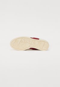 Baskets rouges avec une semelle en caoutchouc blanc texturé. Présente des motifs géométriques en relief et des détails circulaires sur la semelle.