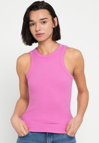 Ροζ εφαρμοστό αμάνικο τοπ με ριμπ υφή και ψηλό λαιμό. Το μοντέλο παρουσιάζεται με τα χέρια στις τσέπες, φορώντας μπλε τζιν παντελόνι.