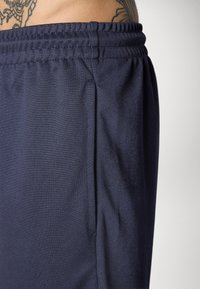 Nahaufnahme einer Person, die dunkelblaue elastische Hosen mit einem Seitentasche trägt und einen Teil eines tätowierten unteren Rückens zeigt.