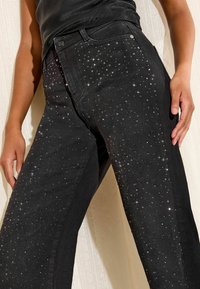 Melni džinsi ar izkliedētām mazām mirdzošām sniegpārsliņām, ko valkā persona, kura stāv pie viegli teksturētas sienas.