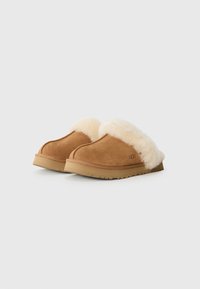 Brune semskede tøfler med en cremefarget pelskrage, flat gummisåle og UGG-logo preget på siden. Myk tekstur, slip-on design.