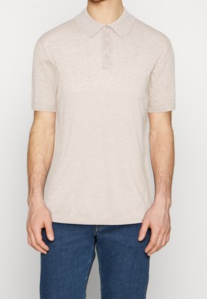 Beżowa koszulka polo z krótkim rękawem o miękkiej fakturze, z guzikowym zapięciem i klasycznym kołnierzykiem. Noszona z ciemnoniebieskimi dżinsami.