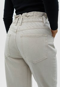 Personne portant un pantalon beige taille haute avec ceinture élastique et poches arrière, mains dans les poches latérales, haut noir à manches longues.